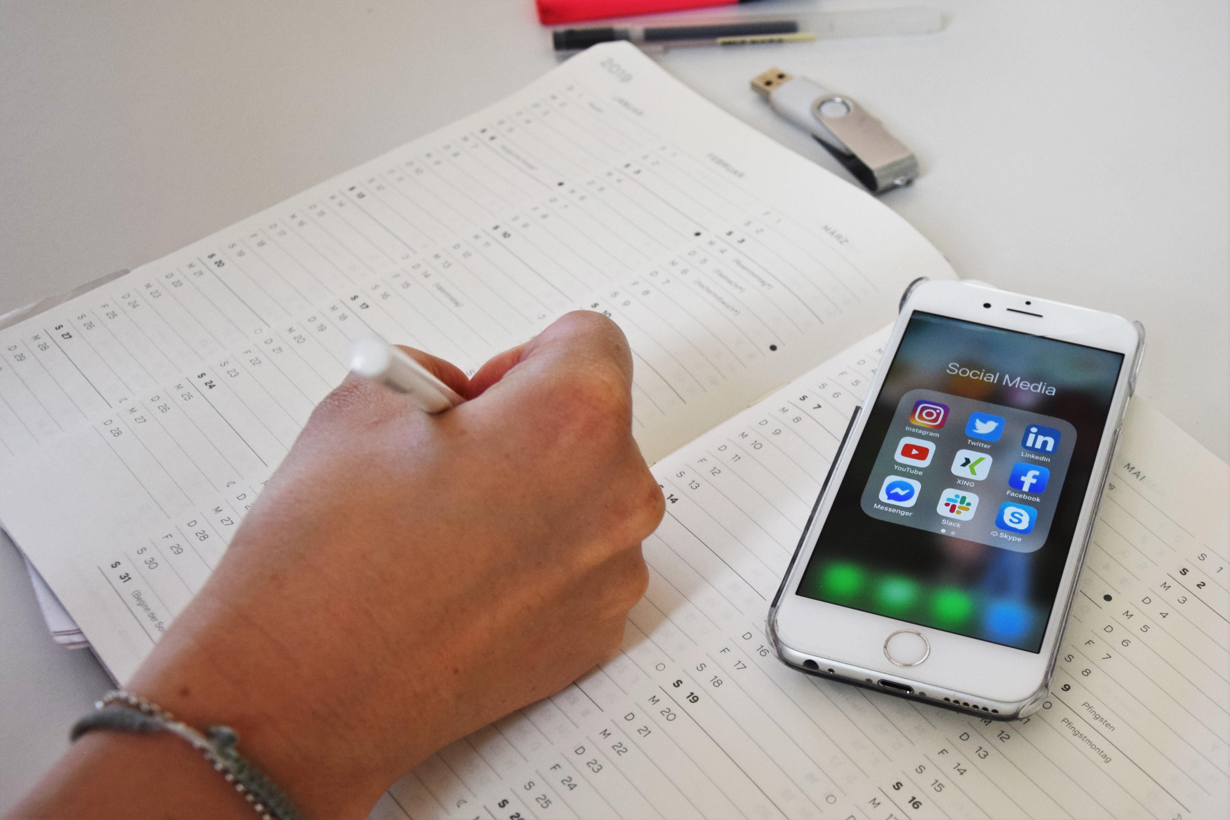 Eine Person trägt einen Termin in einen geöffneten Kalender ein, während auf dem Smartphone daneben Social Media Apps zu sehen sind.